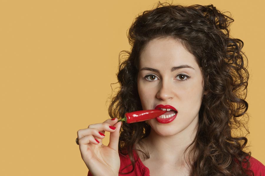 Portrait of a young woman biting red chili pepper over colored background